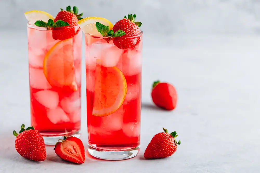 Two tall glasses of strawberry lemonade with fresh strawberries, lemon slices, and mint leaves on a light background.