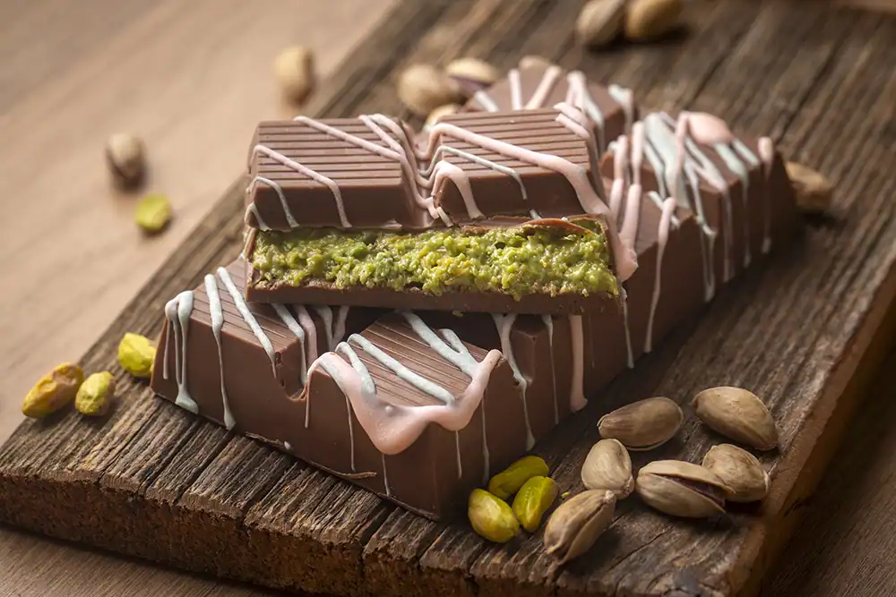 Chocolate bar with pistachio filling on wooden plank surrounded by pistachios.