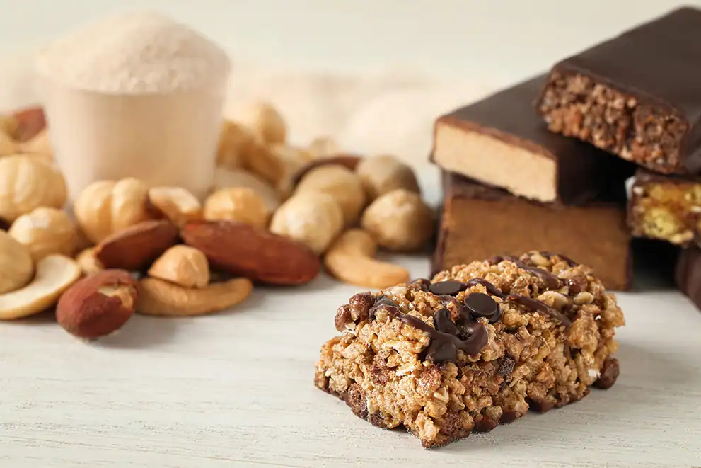Close-up of a protein bar with nuts and chocolate chips, with a bowl of protein powder and assorted nuts in the background.